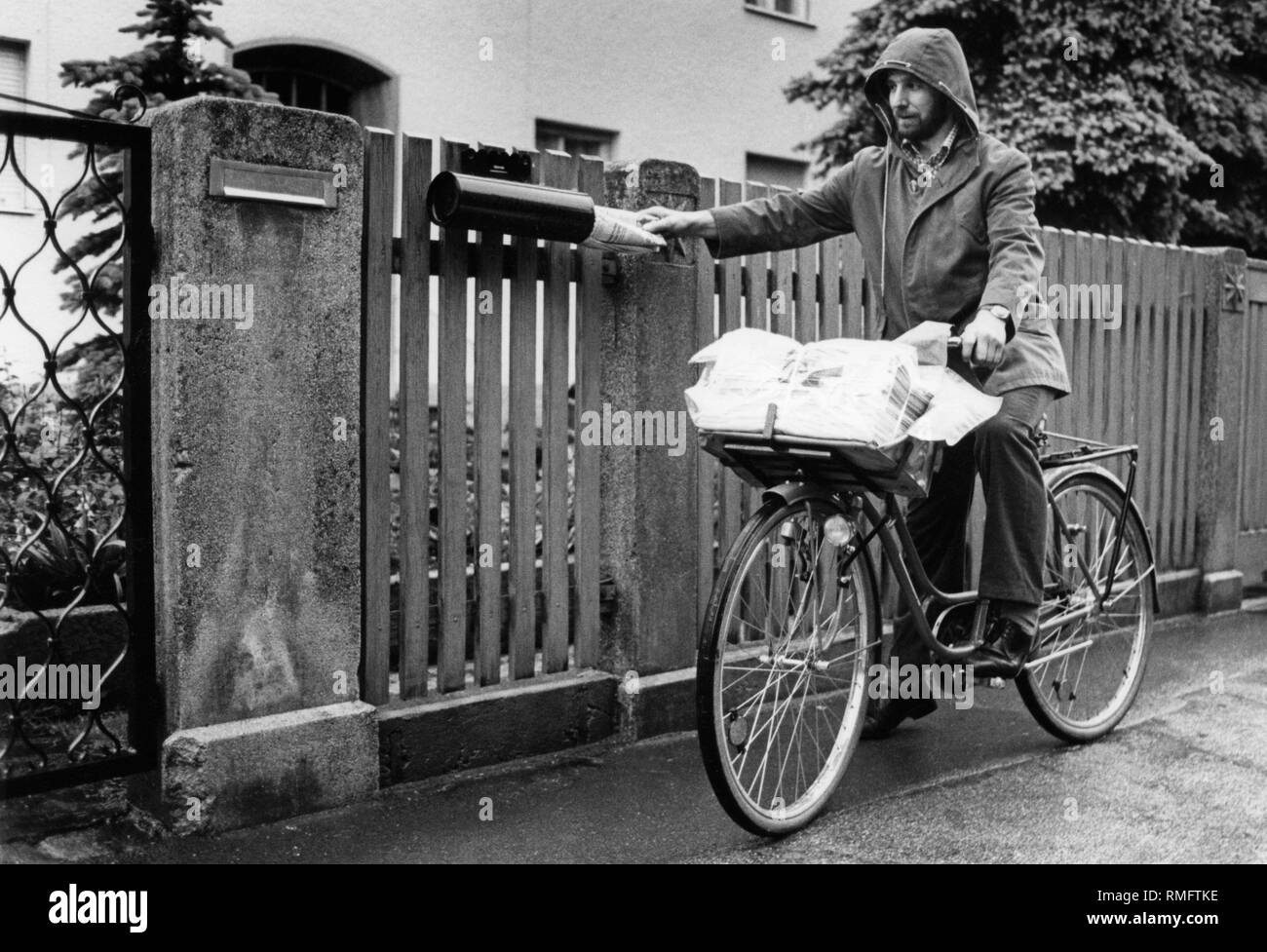 a-newspaper-delivery-person-with-bicycle-and-raincoat-puts-a-newspaper-in-a-mailbox-undated-picture-RMFTKE